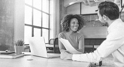 Woman meeting with a man and showing him a piece of paper while she sits behind a laptop.